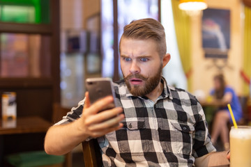 Shocked young hipster man drinking cocoa from a straw and looking at messages in social networks while sitting at a table in a cafe. The concept of chatting and social networks.