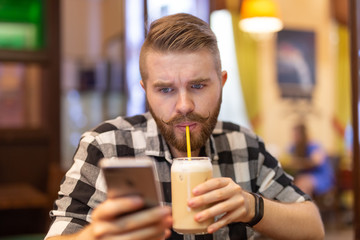 Thoughtful young stylish young hipster man with mustache and beard is looking at social network in smartphone using high-speed free internet. Concept of communication in social networks.