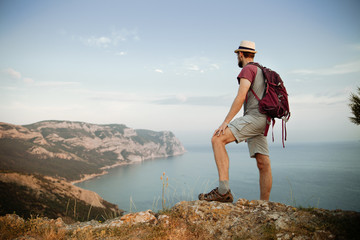 The traveler stands on a stone and looks at the mountains and the sea. Summer hiking concept