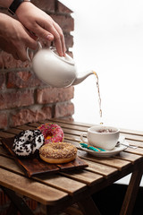 Woman is pouring tea for a tea party with doughnuts: strawberry, oreo and chocolate glazed, served on a brick wall background