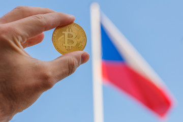 A man fingers are holding a big golden coin of bitcoin in front of Czech Republic flag and blue sky.There is a big golden bitcoin coin on the Czech Republic flag on the background at good weather and