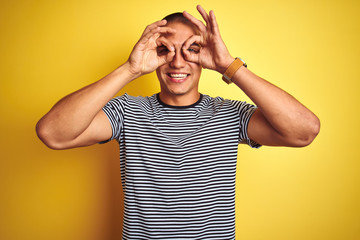 Young handsome man wearing striped t-shirt over yellow isolated background doing ok gesture like binoculars sticking tongue out, eyes looking through fingers. Crazy expression.