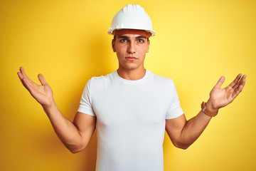 Young handsome man wearing construction helmet over yellow isolated background clueless and confused expression with arms and hands raised. Doubt concept.