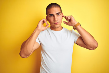 Young caucasian man wearing casual white t-shirt over yellow isolated background covering ears with fingers with annoyed expression for the noise of loud music. Deaf concept.
