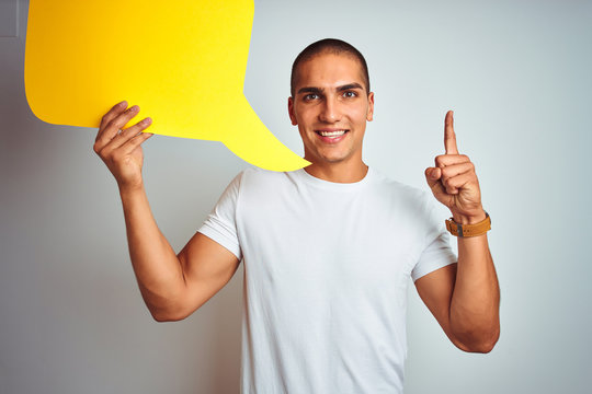 Young man holding yellow speech bubble over white isolated background surprised with an idea or question pointing finger with happy face, number one