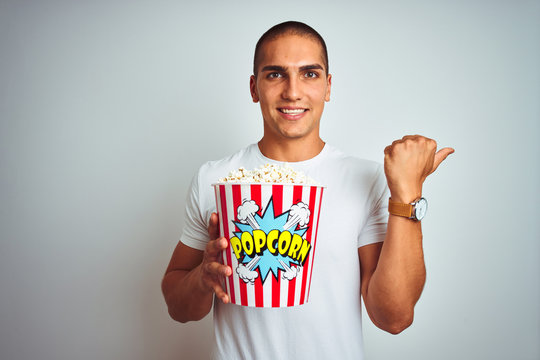 Young handsome man eating popcorn over white isolated background pointing and showing with thumb up to the side with happy face smiling