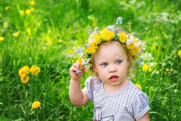 Funny baby girl in a wreath of yellow dandelions on the background of spring grass.