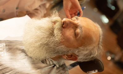 Bearded male sitting in an armchair in a barber shop while hairdresser shaves his beard with a dangerous razor.