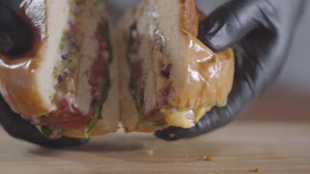 Hands of the chef in black rubber gloves pressing halves of burger and sauce flowing out close-up. Tasty food preparation. Juicy appetizing burger on the table. Slow motion.