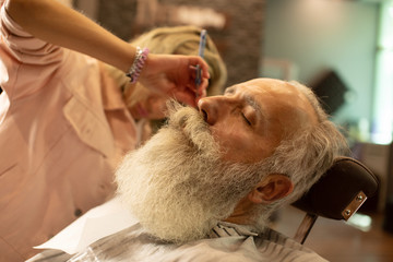 Obraz na płótnie Canvas Bearded male sitting in an armchair in a barber shop while hairdresser shaves his beard with a dangerous razor.