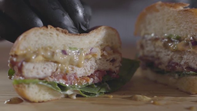 Close-up hands of the chef in black rubber gloves pressing halves of burger and sauce flowing out. Tasty food preparation. Juicy mouth-watering burger on the table. Slow motion.