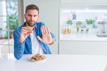Handsome man eating chocolate chips cookies with open hand doing stop sign with serious and confident expression, defense gesture