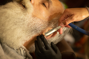 Obraz premium Bearded male sitting in an armchair in a barber shop while hairdresser shaves his beard with a dangerous razor.