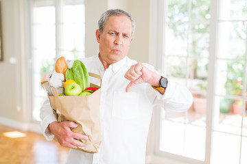 Handsome senior man holding a paper bag of fresh groceries from the supermarket with angry face, negative sign showing dislike with thumbs down, rejection concept