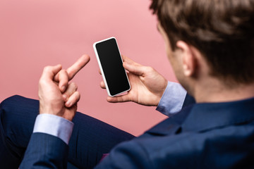 partial view of businessman showing middle finger while holding smartphone with blank screen isolated on pink