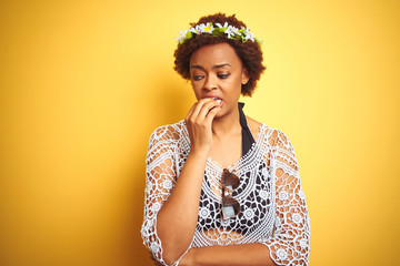 Young african american woman with afro hair wearing flowers crown over yellow isolated background looking stressed and nervous with hands on mouth biting nails. Anxiety problem.