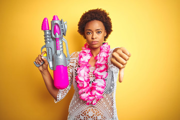 Young african american woman with afro hair wearing flower hawaiian lei and water gun with angry face, negative sign showing dislike with thumbs down, rejection concept
