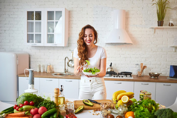 Young happy woman eating salad in the beautiful kitchen with green fresh ingredients indoors. Healthy food concept