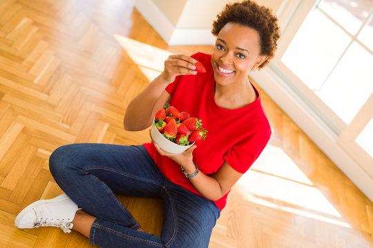 Beautiful young african woman with afro hair eating fresh strawberries sitting on the floor