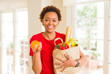 Young beautiful african american woman holding paper bag full of fresh healthy groceries and picking vegetables