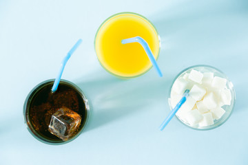Excessive sugar consumption concept - cola, juice and sugar cubes in glasses on blue background, copy space