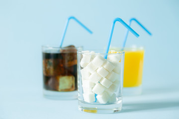 Excessive sugar consumption concept - cola, juice and sugar cubes in glasses on blue background,...