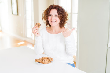 Senior woman eating chocolate chips cookies at home pointing and showing with thumb up to the side with happy face smiling