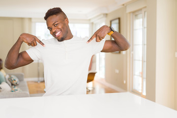 Handsome african american man on white table at home looking confident with smile on face, pointing...
