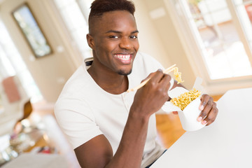 Handsome african man eating asian noodles in a delivery box, smiling enjoying lunch using chopsticks