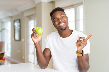 Young african american man eating fresh green apple very happy pointing with hand and finger to the side