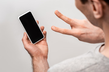 cropped veiw of man showing victory sign while holding smartphone with blank screen isolated on grey