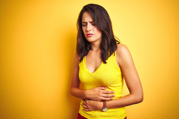 Young beautiful woman wearing t-shirt standing over yellow isolated background with hand on stomach because nausea, painful disease feeling unwell. Ache concept.