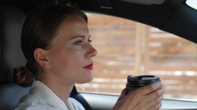 Close-up portrait of confident woman in business clothes drinking morning coffee while driving car through cottage town. Formally dressed woman enjoying coffee drink going to work in automobile
