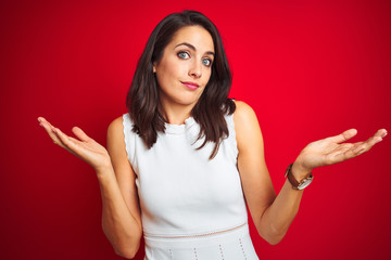 Young beautiful woman wearing white dress standing over red isolated background clueless and confused expression with arms and hands raised. Doubt concept.