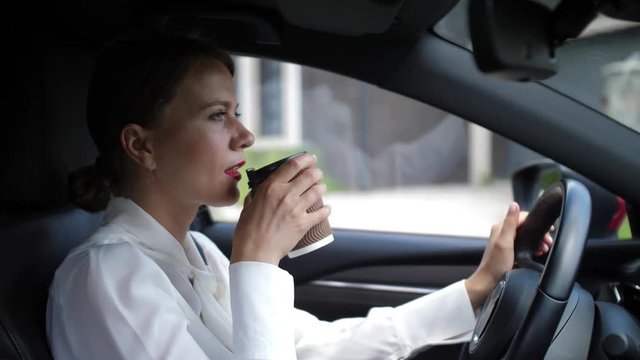 Close-up of confident female in business clothes drinking coffee with pleasure while driving car through cottage town. Formally dressed businesswoman enjoying morning coffee going to work by own car