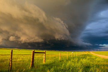 A dark shelf cloud and storm approach as the sun shines brightly looking down a fence in the rural countryside.