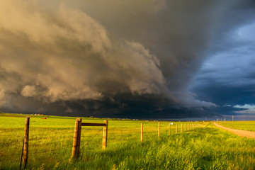 A dark shelf cloud and storm approach as the sun shines brightly looking down a fence in the rural...