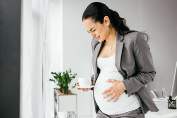 pregnant woman standing in office with cup of coffe and enduring pain