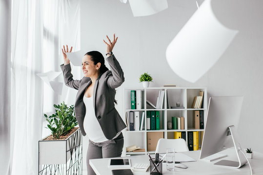 Irritated Pregnant Woman Throwing Papers In Air While Standing Near Table In Office