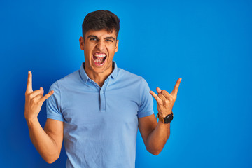 Young indian man wearing casual polo standing over isolated blue background shouting with crazy...