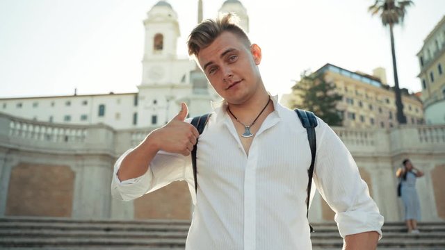 Brutal handsome young boy looking confidently on camera and smiling slightly. Man meeting wonderful sunrise in the city on the stairs and showing his thumb up. Close-up portrait of brutal tourist man