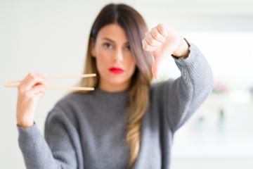 Young beautiful woman holding asian chopsticks at home with angry face, negative sign showing...