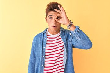 Young handsome man wearing striped t-shirt and denim shirt over isolated yellow background doing ok gesture shocked with surprised face, eye looking through fingers. Unbelieving expression.