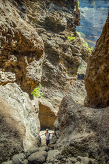 Young traveler stays on the top of huge boulder in the Masca gorge, Tenerife, showing solidified volcanic lava flow layers and arch formation. Ravine leads down to the ocean from a 900m altitude
