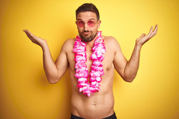 Young handsome shirtless man wearing sunglasses and pink hawaiian lei over yellow background...