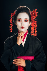 beautiful geisha in black and red kimono and flowers in hair with hand near chin isolated on black