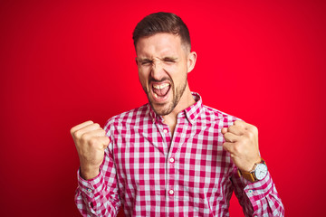 Naklejka premium Young handsome man over red isolated background very happy and excited doing winner gesture with arms raised, smiling and screaming for success. Celebration concept.
