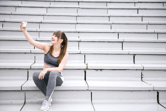 Young woman in sportswear sitting on concrete bleachers and making selfie