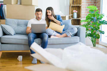 Obraz premium Young beautiful couple relaxing sitting on the sofa of new house using computer laptop scared in shock with a surprise face, afraid and excited with fear expression