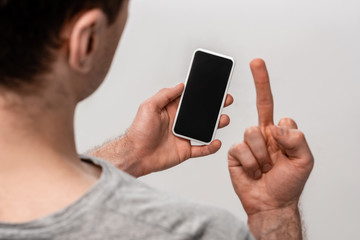 copped view of man showing middle finger while holding smartphone with blank screen isolated on grey